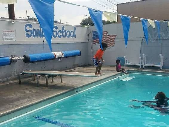 Children playing at a swim school, with one preparing to jump from a diving board into a clear blue pool.