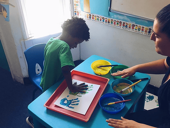 A child creates handprints on paper at a desk, with an adult gesturing nearby and several bowls of colorful paint.