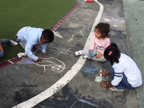 Three children sit on the ground drawing with colorful chalk on a concrete walkway next to a grassy area.