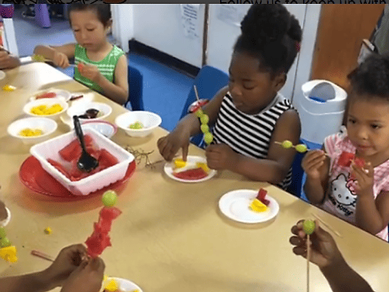 Children sitting at a table making fruit skewers with watermelon, grapes, and pineapple.