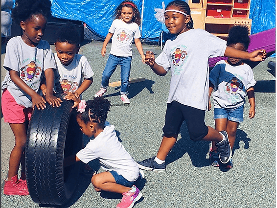 Children in matching graphic t-shirts play with a large tire on a paved playground.