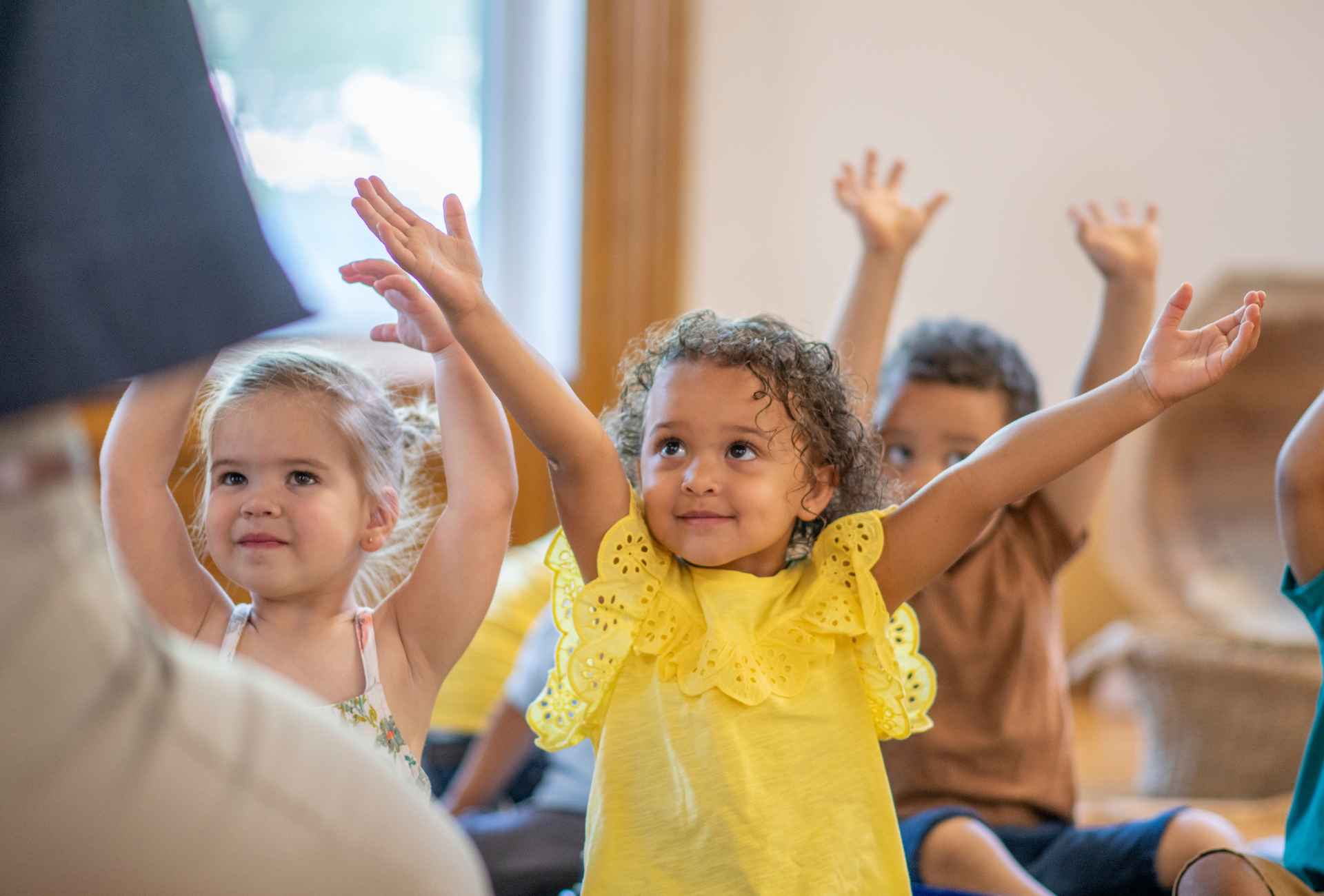 Children sitting indoors with arms raised, smiling and participating in a group activity.