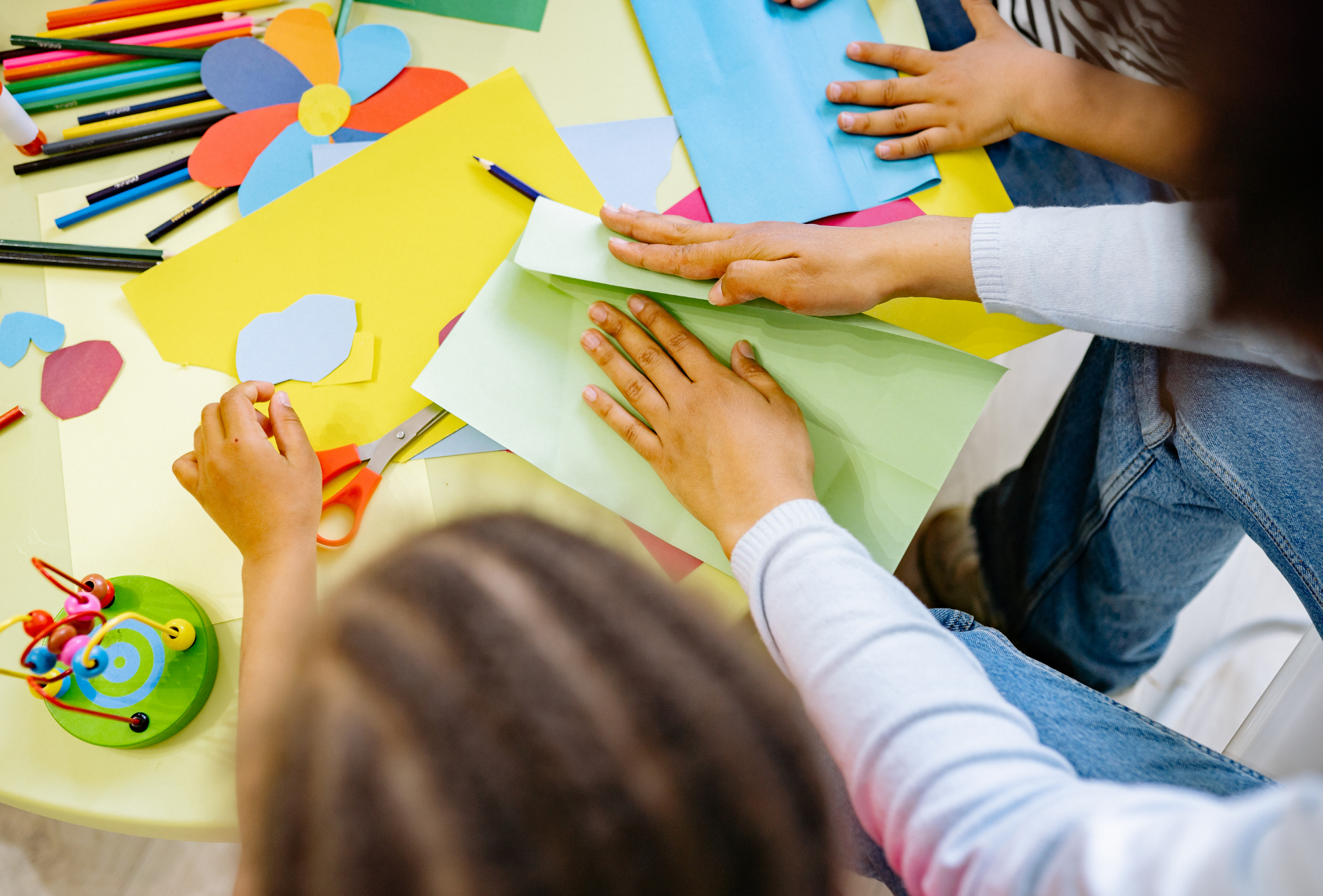 Hands fold a light green piece of paper on a table cluttered with colorful craft supplies and construction paper.