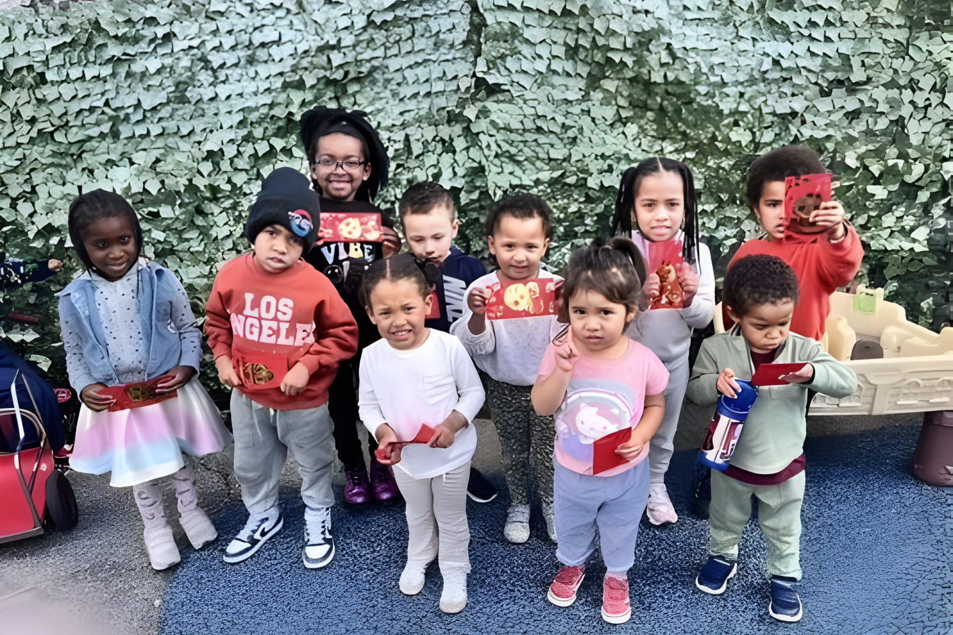 A group of children stand outside against a leaf-covered wall, smiling and holding small red cards.