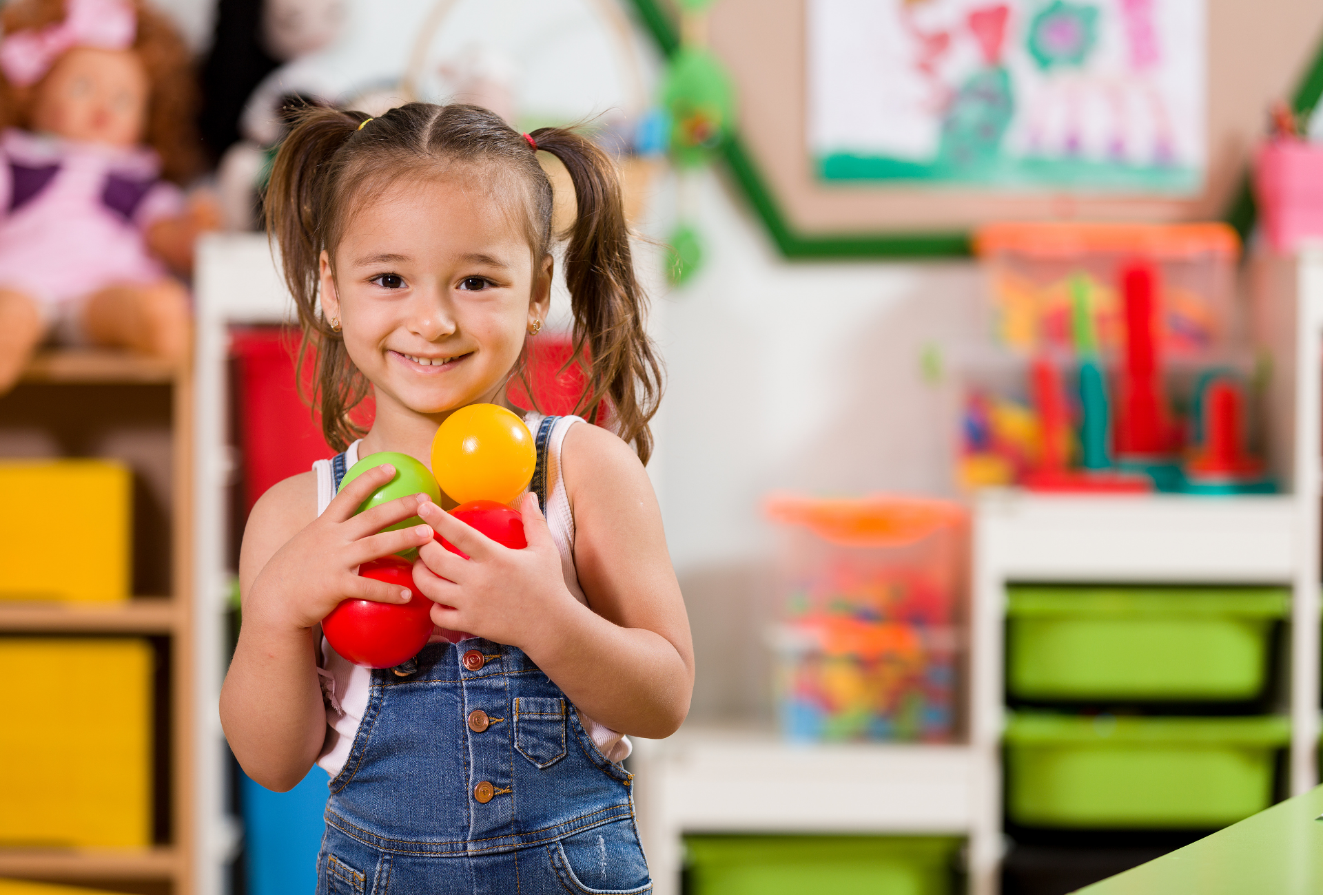 A smiling child in denim overalls holds three colorful toy balls in a playroom filled with shelves and storage bins.