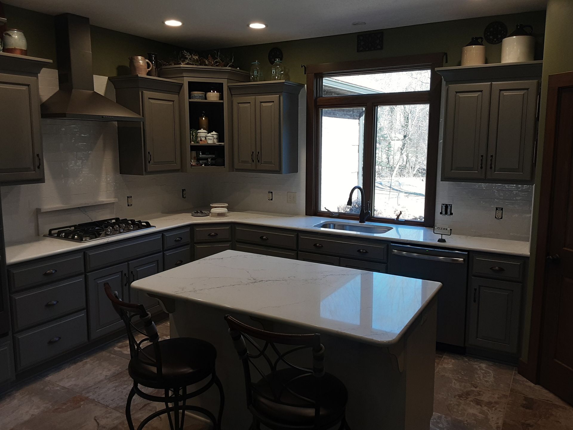 U-shaped kitchen with gray cabinets, white countertops, island with stools, and window overlooking outdoors.