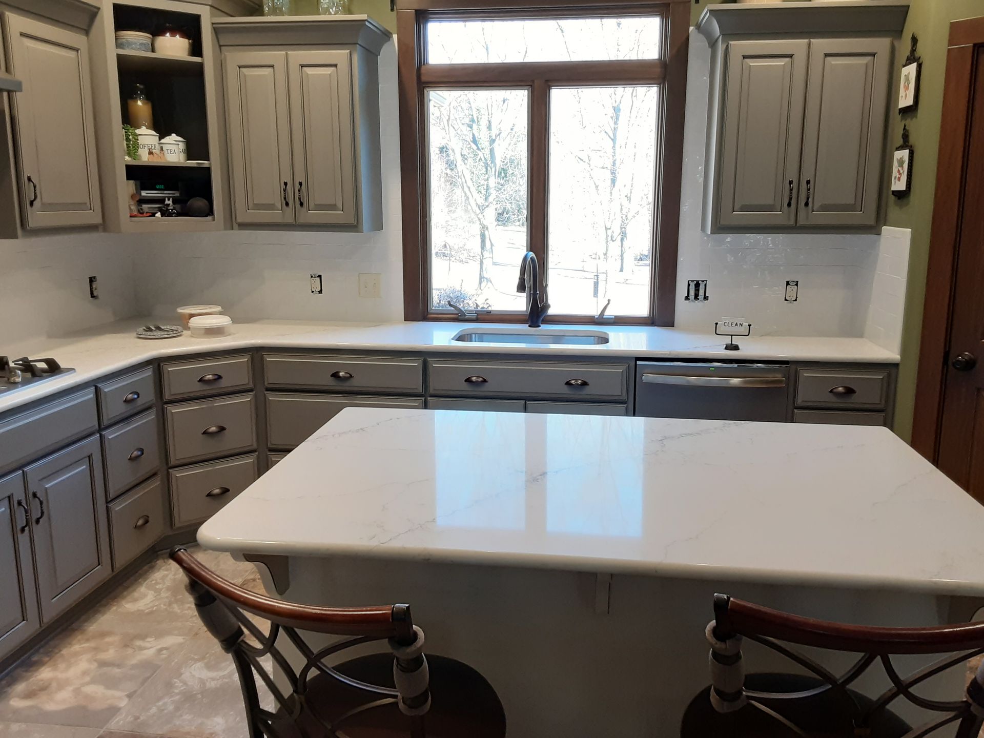 Gray kitchen cabinets with white countertops, a sink, and an island with stools.