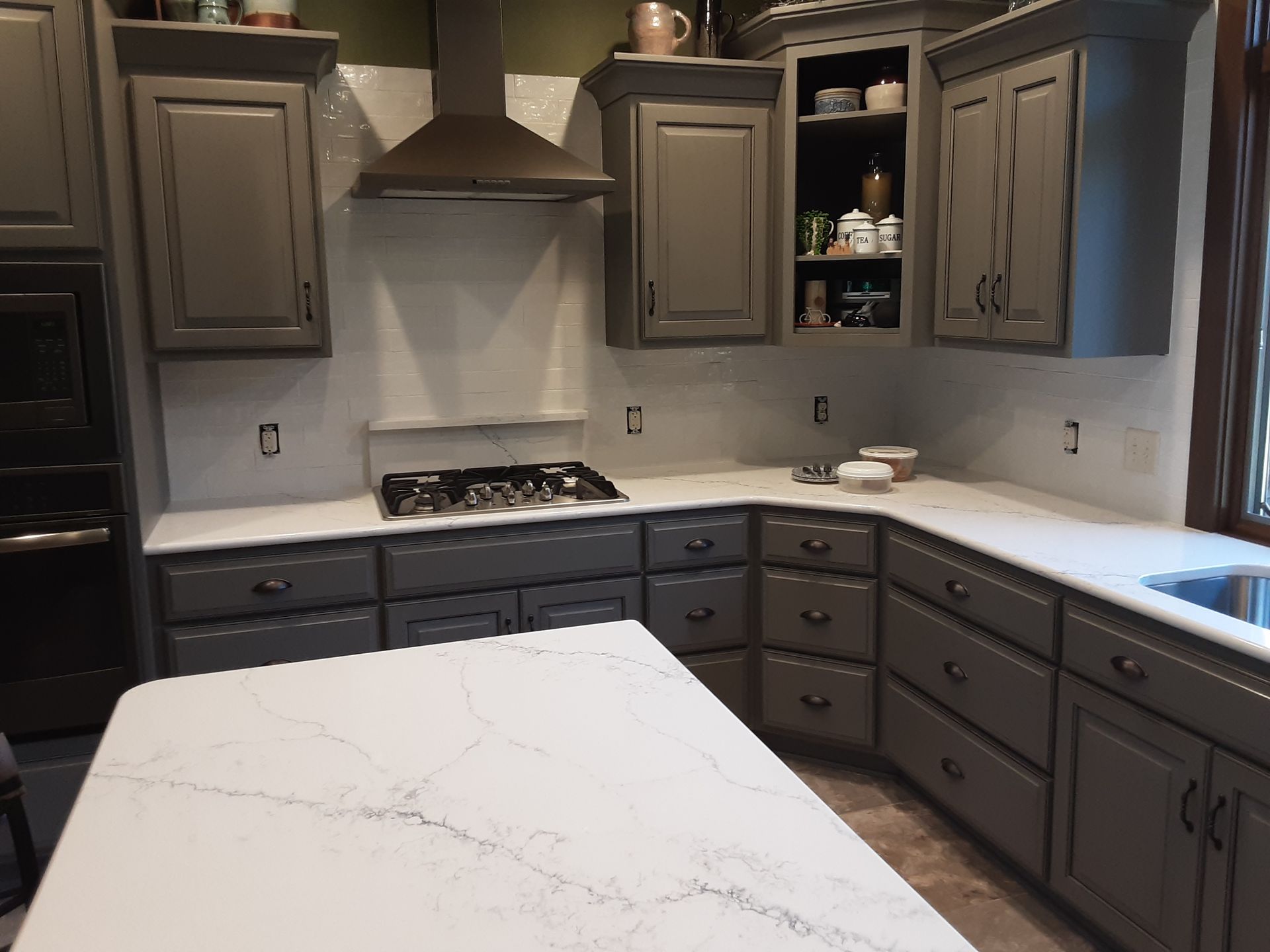 Gray kitchen cabinets with white countertops, cooktop, and range hood.