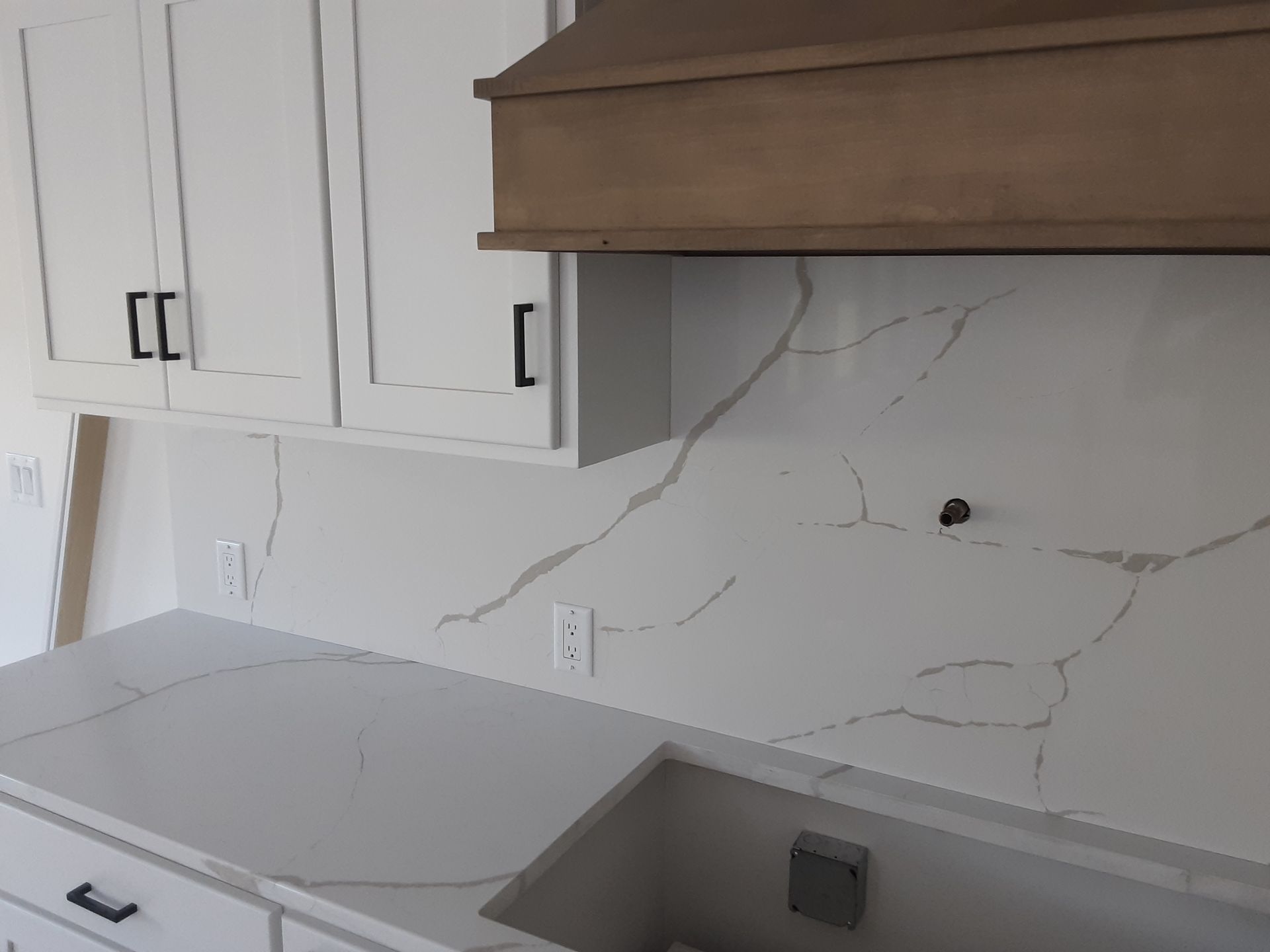 Kitchen with white cabinets, countertops, and backsplash featuring grey veining; wooden range hood.