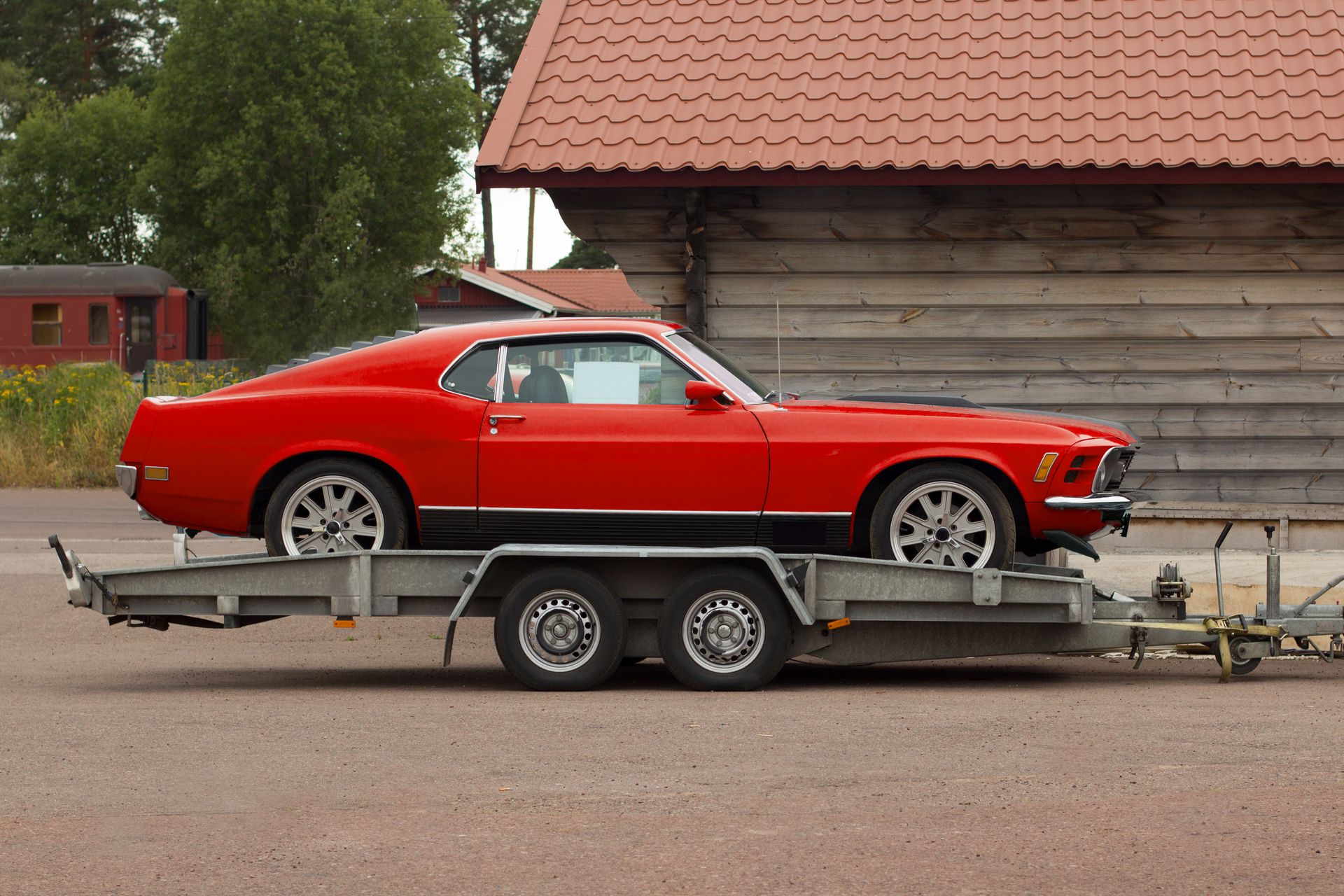 A red mustang is on a trailer in front of a wooden building