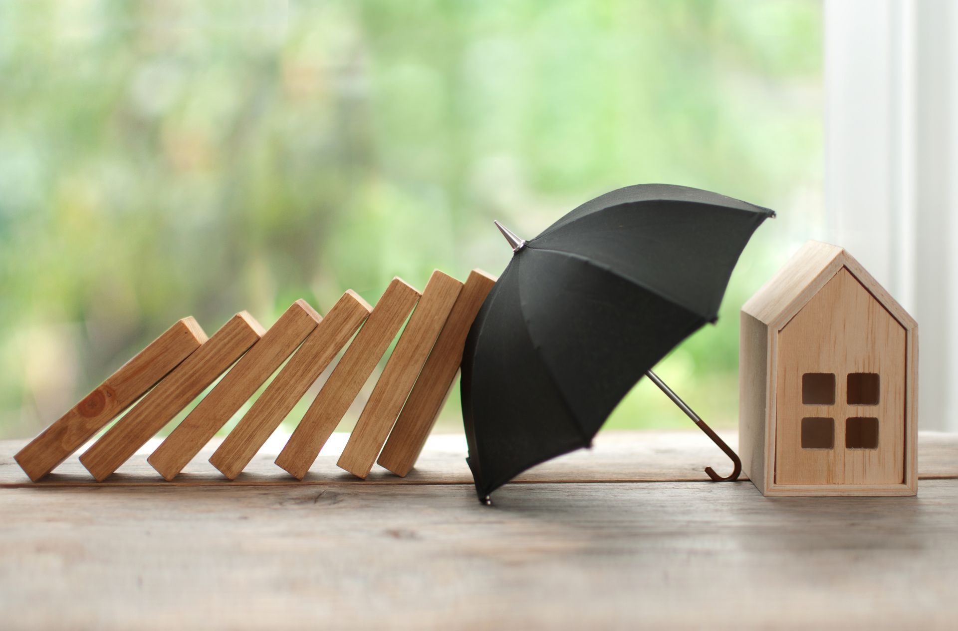 A black umbrella is protecting a wooden house from falling dominoes.
