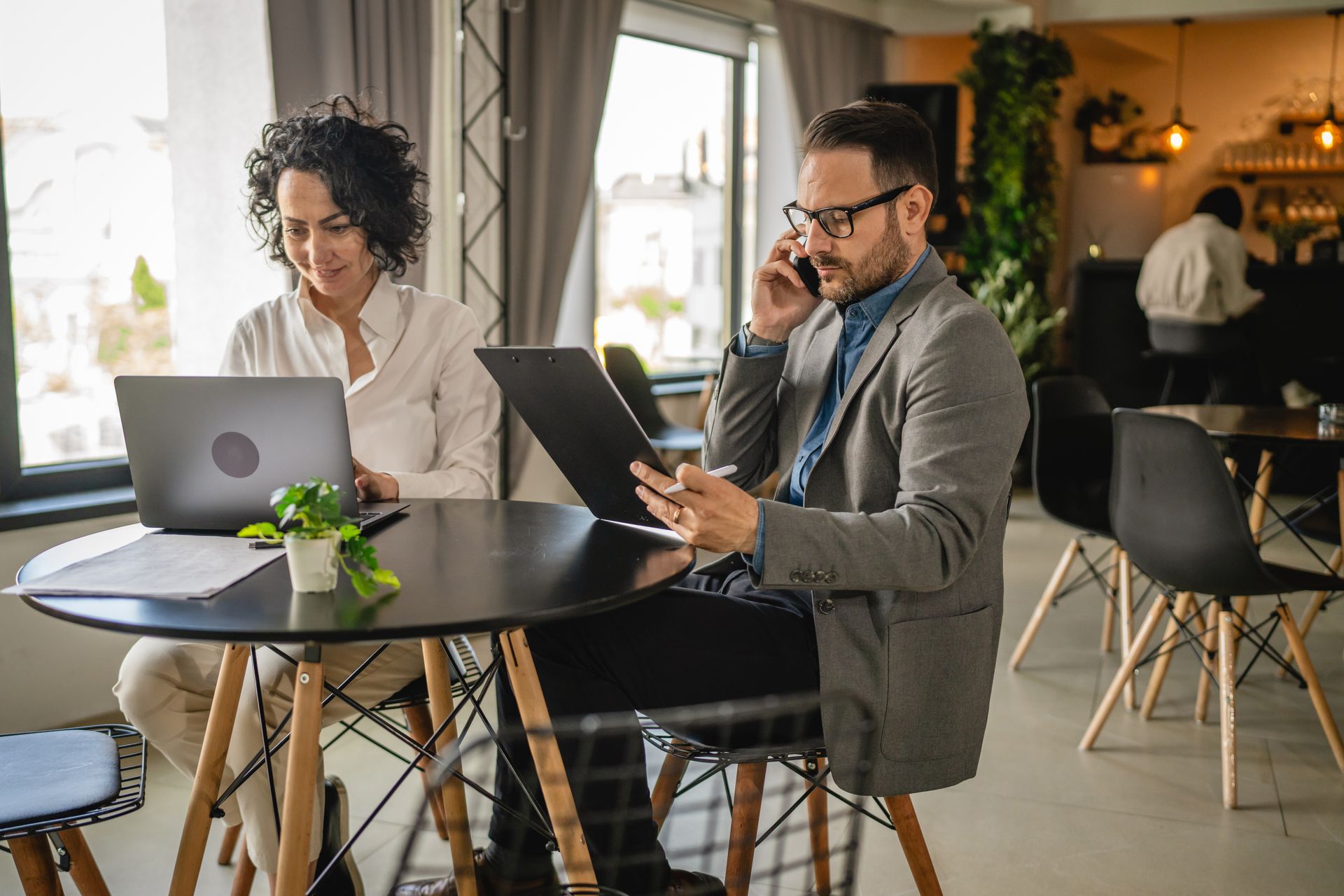 A man and a woman are sitting at a table with laptops and talking on cell phones.