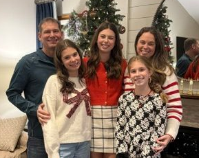 Five smiling people pose indoors by a decorated Christmas tree, wearing festive winter sweaters.