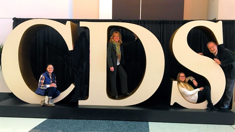 People posing inside large beige “CDS” letters in a bright indoor lobby.