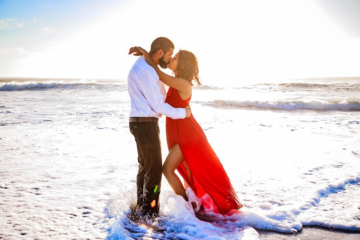 Ein Mann und eine Frau küssen sich am Strand. Die Frau trägt ein rotes Kleid.