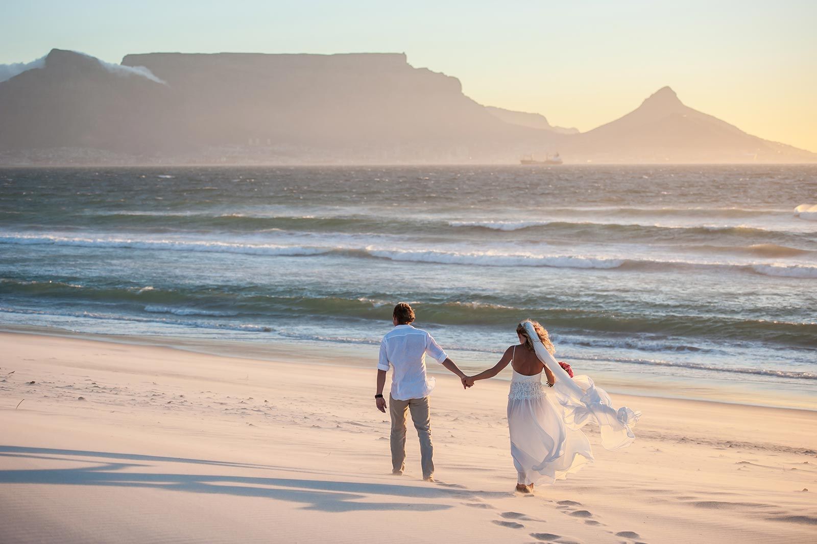 A bride and groom are walking on the beach holding hands.