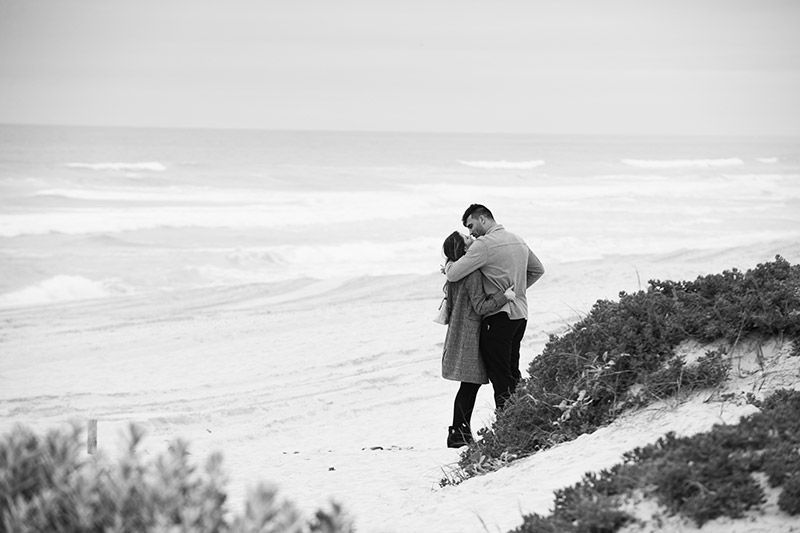 A black and white photo of a man and woman hugging on a beach.