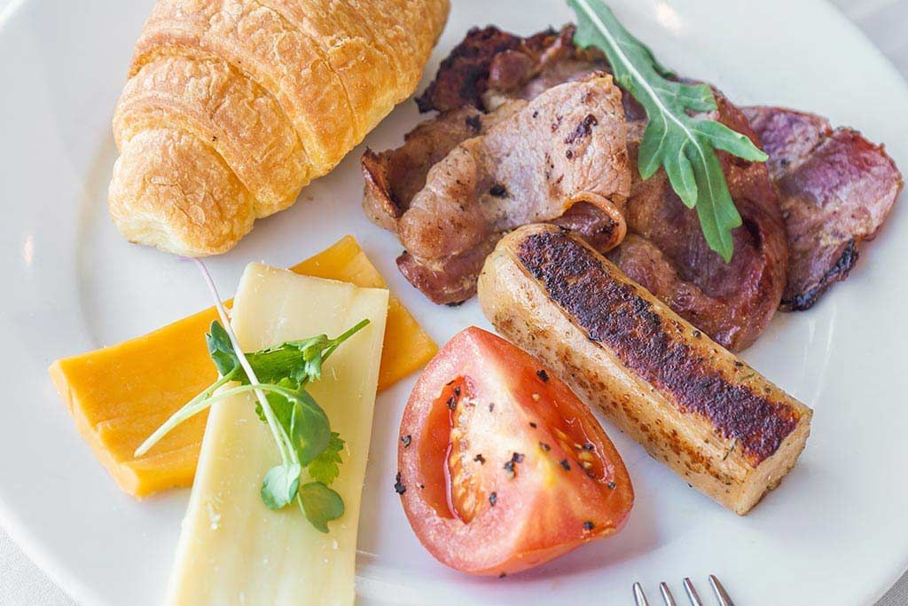 Un plato de comida con un croissant, queso, tocino, tomates y salchichas.