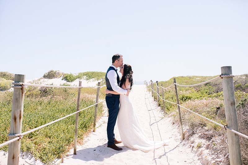 A bride and groom are kissing on a sandy path.