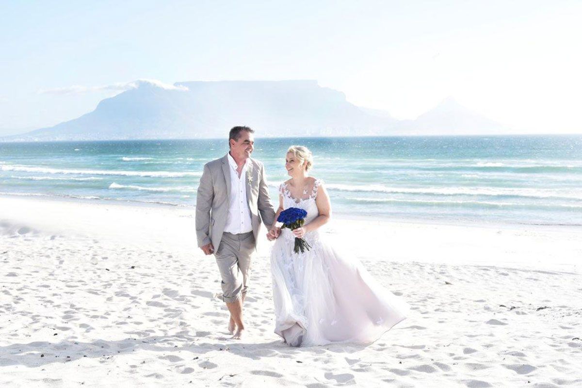 A bride and groom are walking on the beach holding hands.