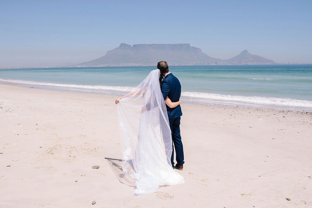 A bride and groom are kissing on the beach with a mountain in the background.