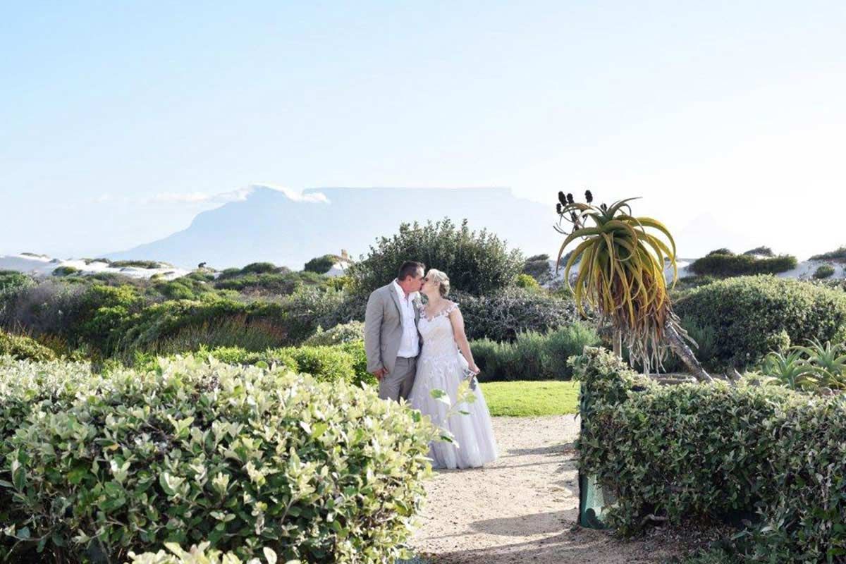A bride and groom are kissing in a garden with a mountain in the background.