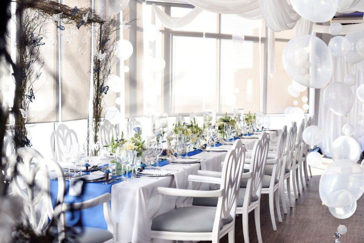 A long table with a blue table cloth and white chairs in a room with balloons hanging from the ceiling.