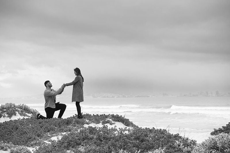 Ein Mann kniet nieder, um einer Frau am Strand einen Heiratsantrag zu machen.