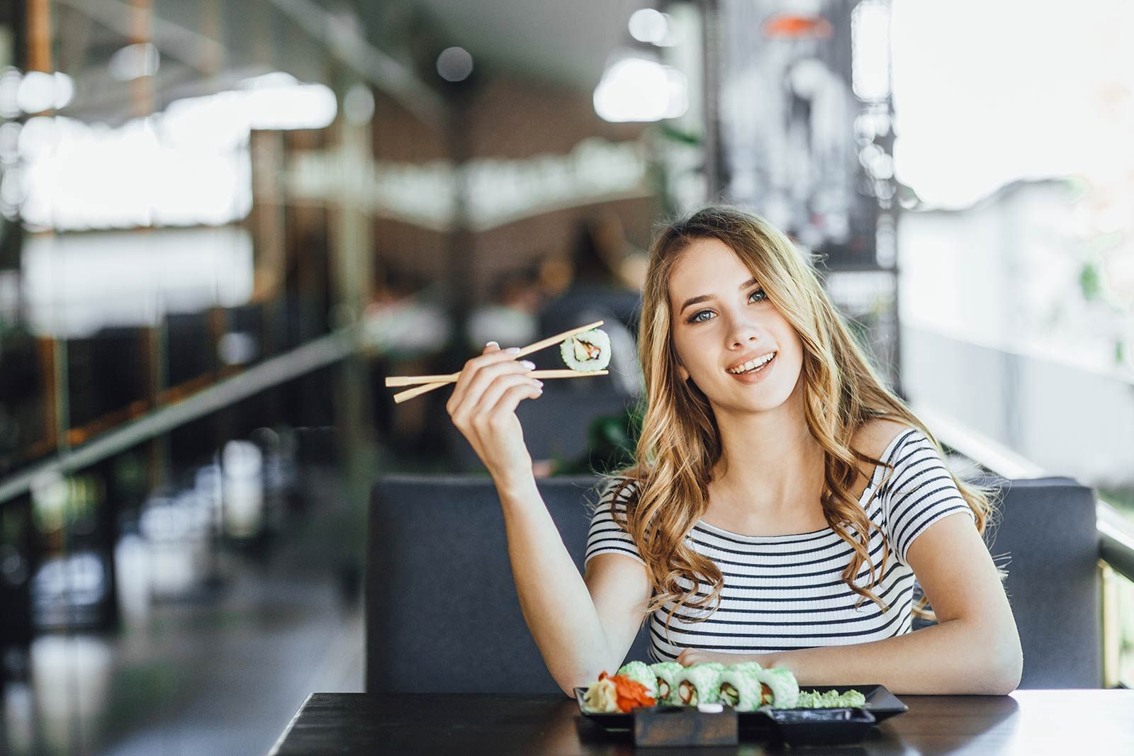 A woman is sitting at a table eating sushi with chopsticks.