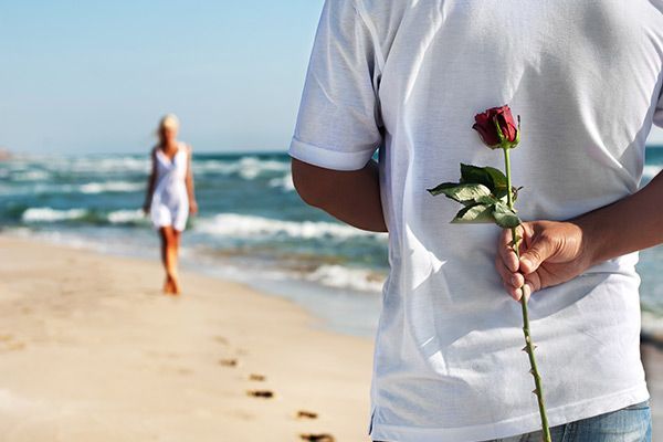 Een man houdt een roos achter zijn rug vast op het strand.