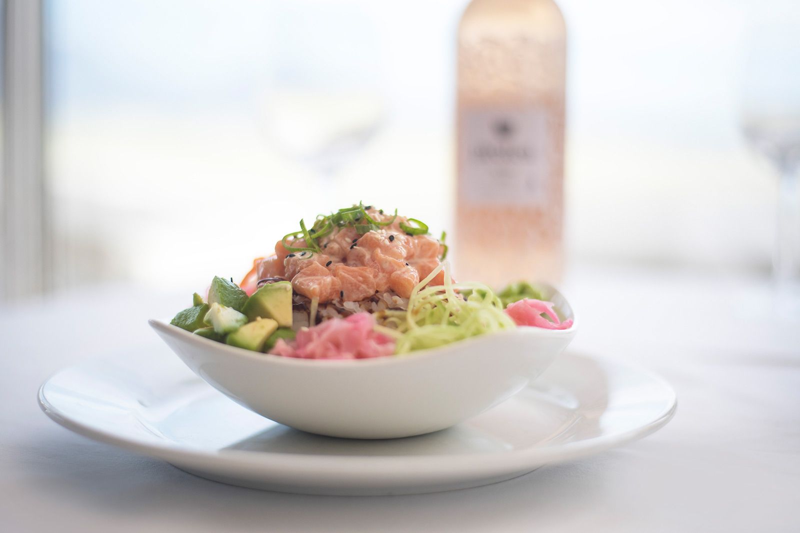 A bowl of food on a plate with a bottle of wine in the background.