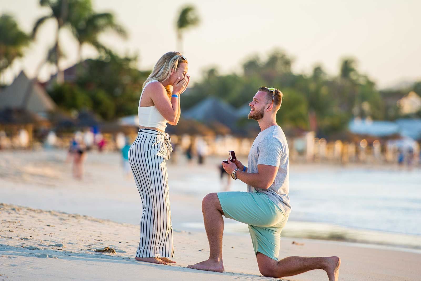 A man is kneeling down on the beach to propose to a woman.