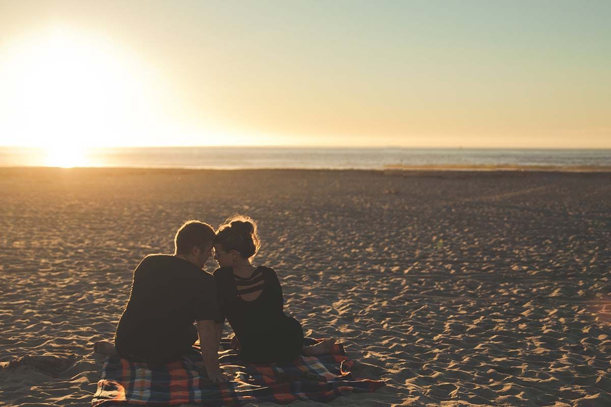 Un uomo e una donna sono seduti su una coperta sulla spiaggia al tramonto.