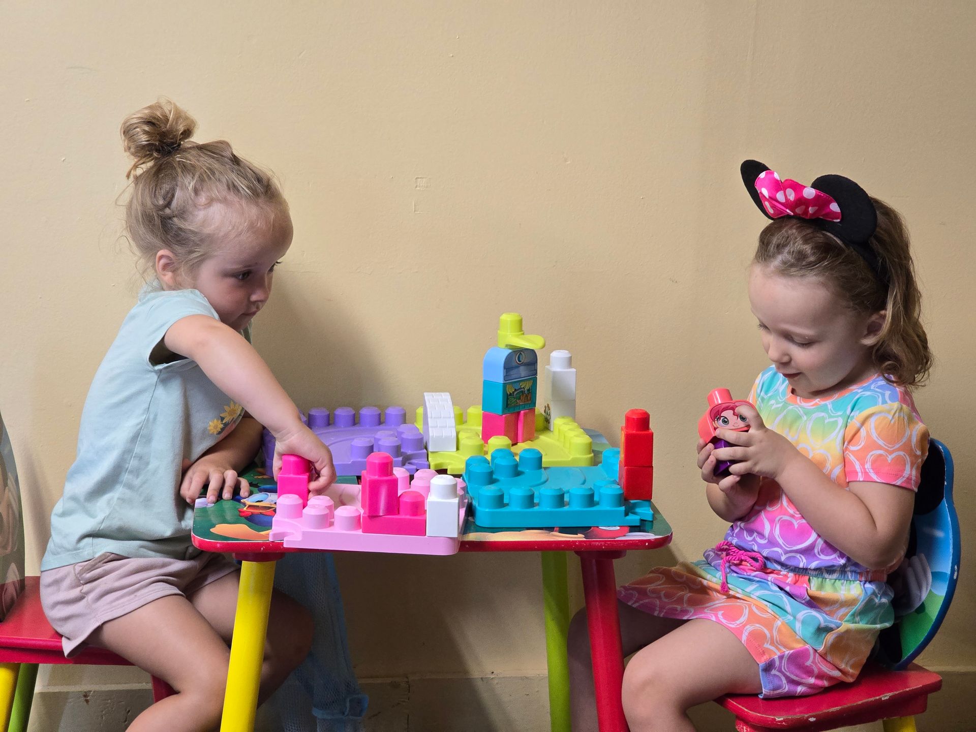 Two young girls building with blocks at a colorful table. One wears a Minnie Mouse headband.