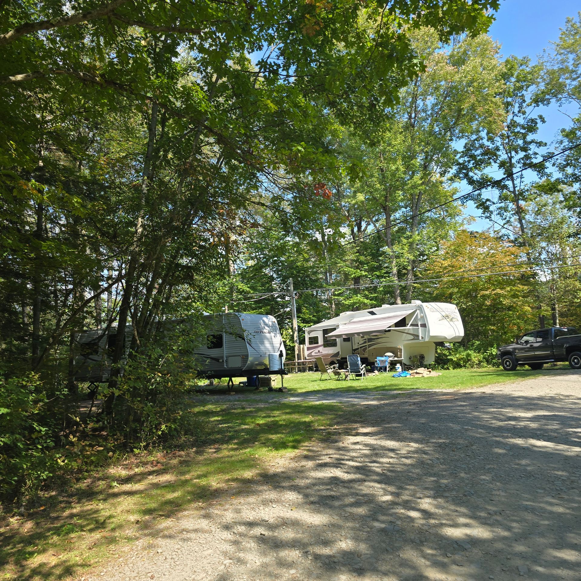 Campground scene: RVs and campers parked in a grassy area, surrounded by trees. People are sitting at a picnic table.