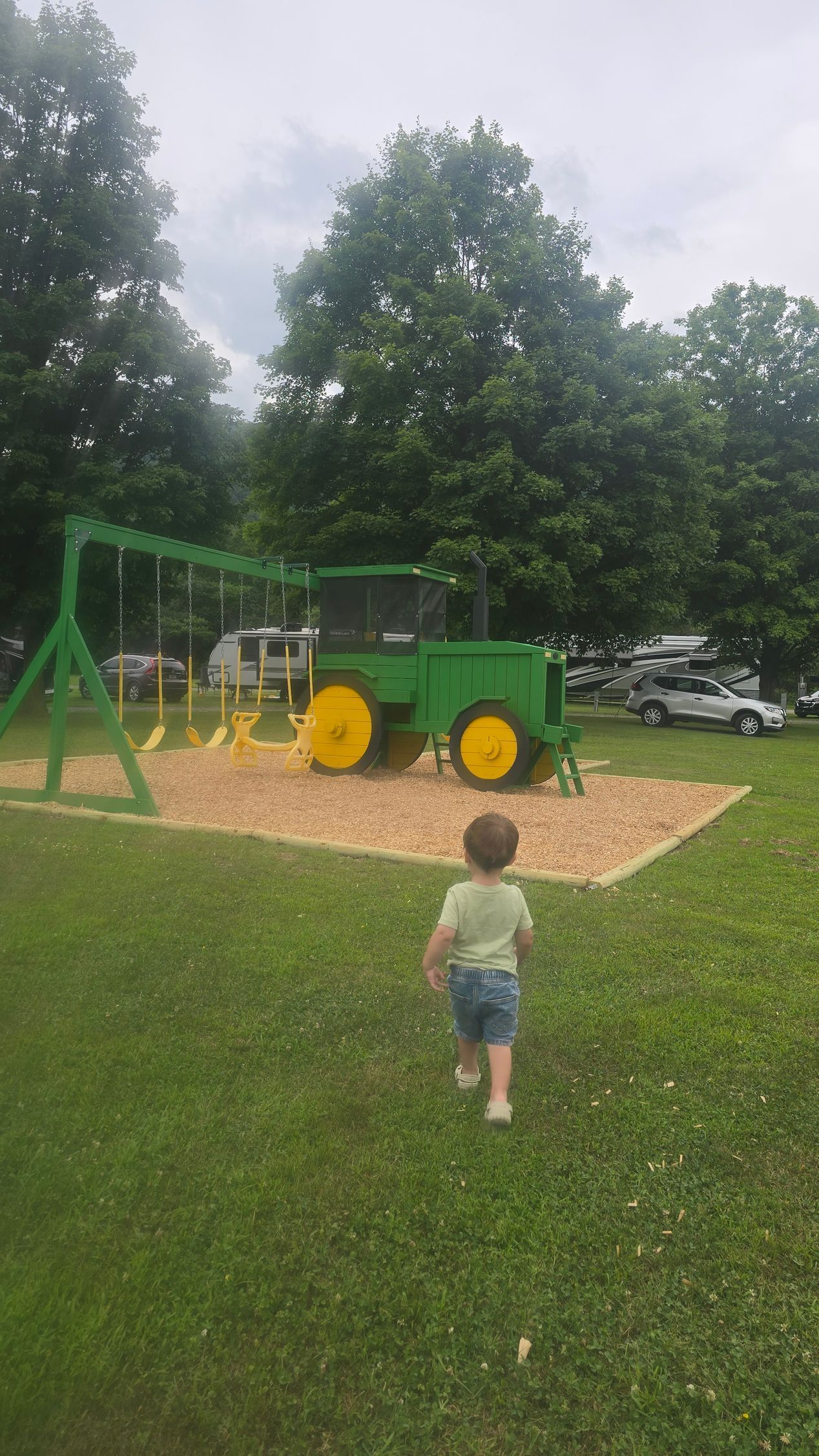 A child runs toward a John Deere tractor-themed playground. Green and yellow, grassy outdoor setting.