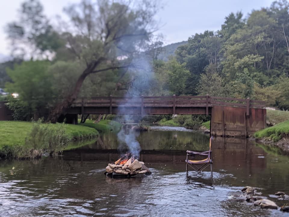 Campfire in a river with an empty chair nearby and a bridge in the background.