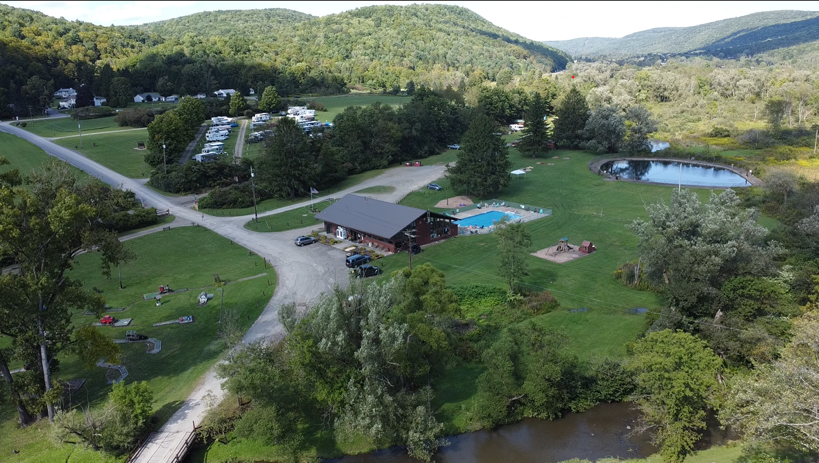 Campground with a cabin, pool, pond, and tents, nestled in a green valley under a sunny sky.