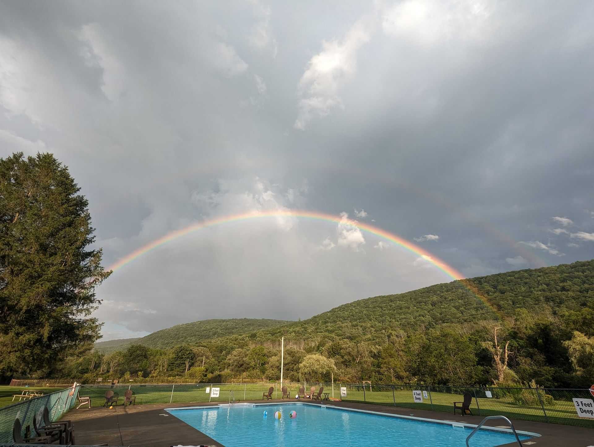 Rainbow arcs over a swimming pool and forested hills under a cloudy sky.