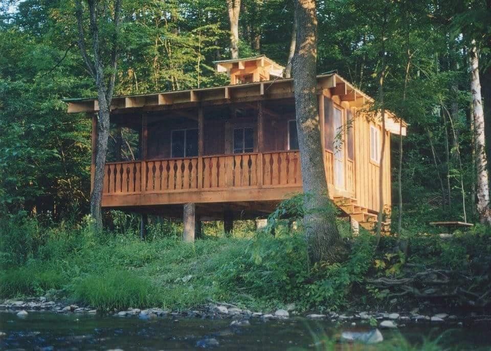 Wooden cabin on stilts in a wooded area, with a porch and small chimney.