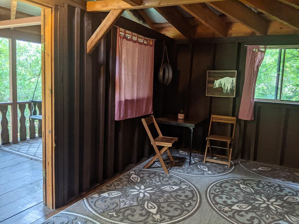 Cozy cabin interior with dark wood walls, a table, chairs, and a decorative rug. Porch visible on the left.