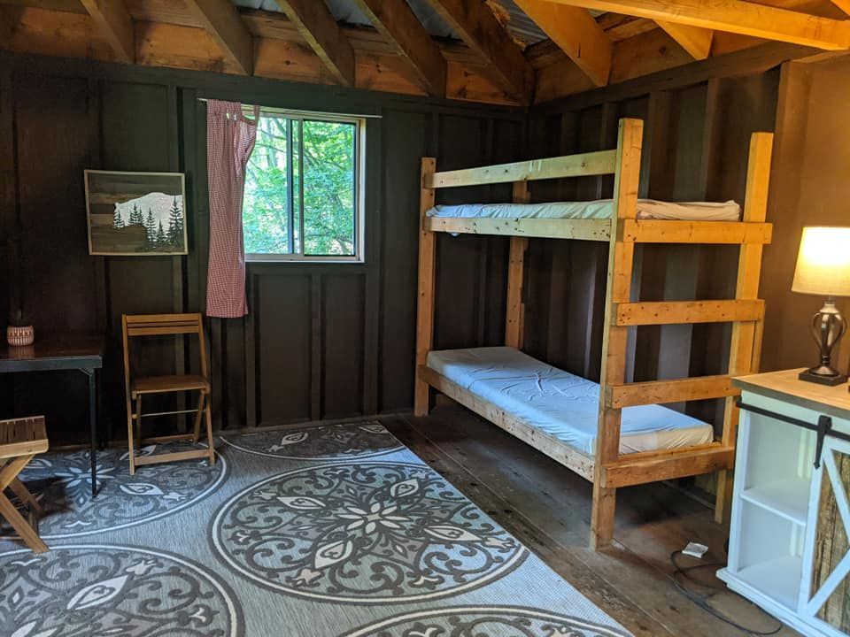 A rustic cabin bedroom with a bunk bed, window, rug, desk, and a lamp.