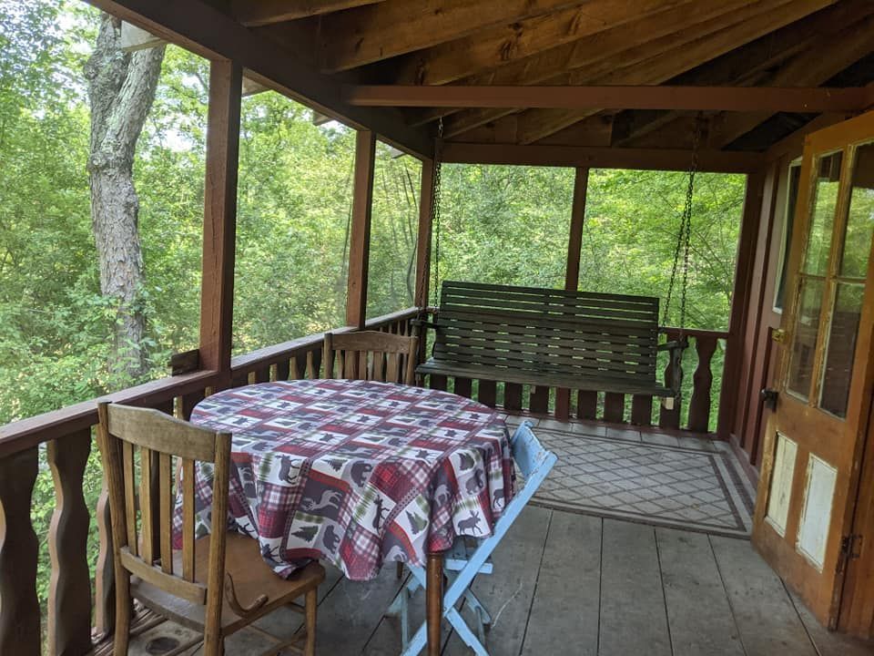 A wooden porch with a table and chairs, a swing, and a view of trees.