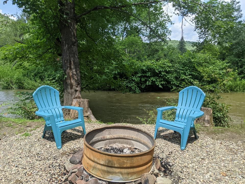 Two blue Adirondack chairs face a fire pit next to a river, with lush green foliage in the background.