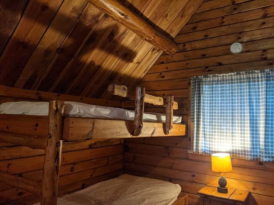 Log cabin interior with bunk beds, small window, and lit bedside lamp.