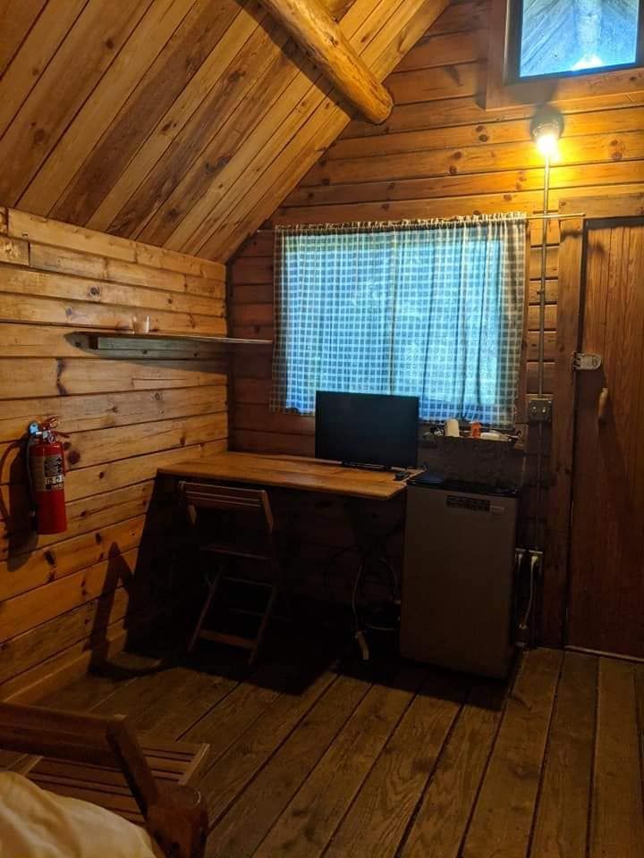 Cabin interior with wooden walls and floors. Desk with TV, window with curtain, small fridge, fire extinguisher.