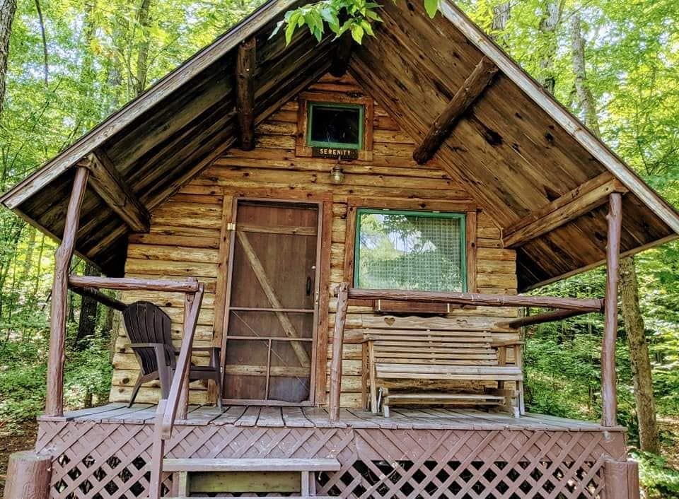 Rustic log cabin with porch, bench, and screen door; surrounded by trees.