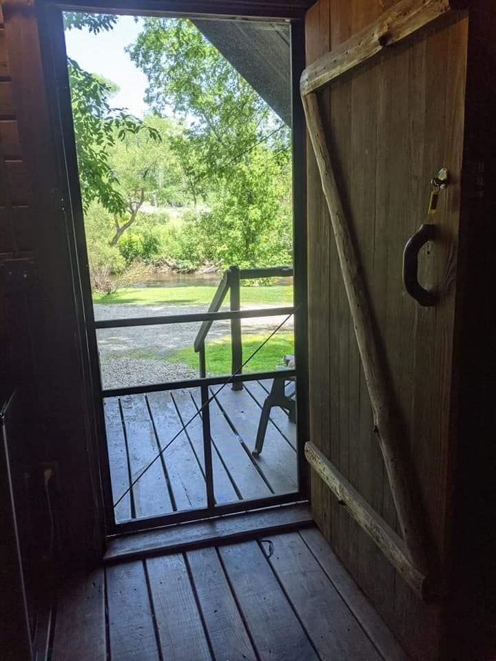 Open wooden door with a view of trees, water, and a railing. Interior is dark, exterior is bright green.