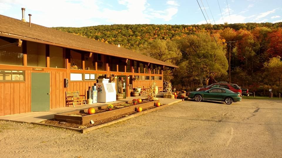 A wooden building with a mountain backdrop and colorful autumn foliage. Pumpkins and parked cars are visible.