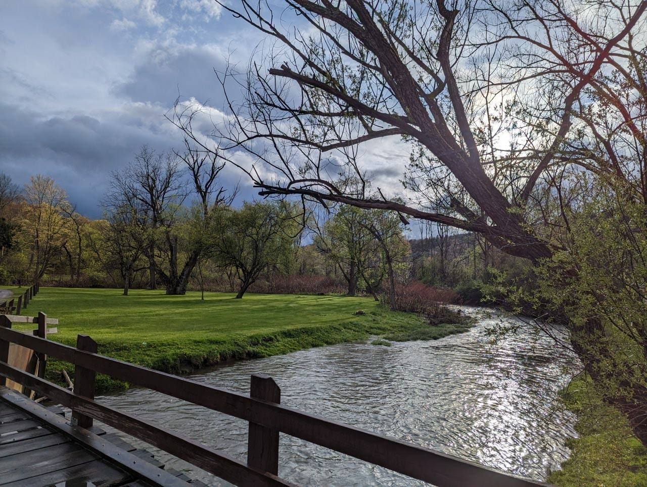 A river winds under a wooden bridge through a grassy field, trees in the background under a cloudy sky.